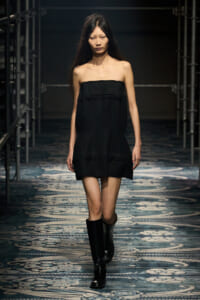 Model walks a runway in a strapless black mini dress with large pom-pom-like buttons and knee-high black boots.