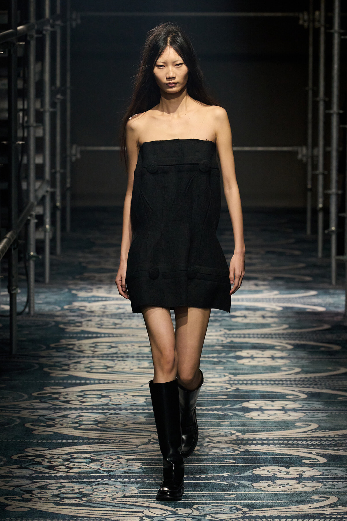 Model walks a runway in a strapless black mini dress with large pom-pom-like buttons and knee-high black boots.