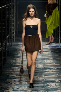 Model walking a fashion runway in a navy strapless knit top with a brown mini skirt, black loafers, and a small brown handbag