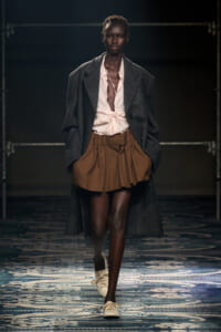 Model walking runway in a dark herringbone overcoat, pink tied blouse, and brown mini skirt; white sneakers visible in a fashion show.