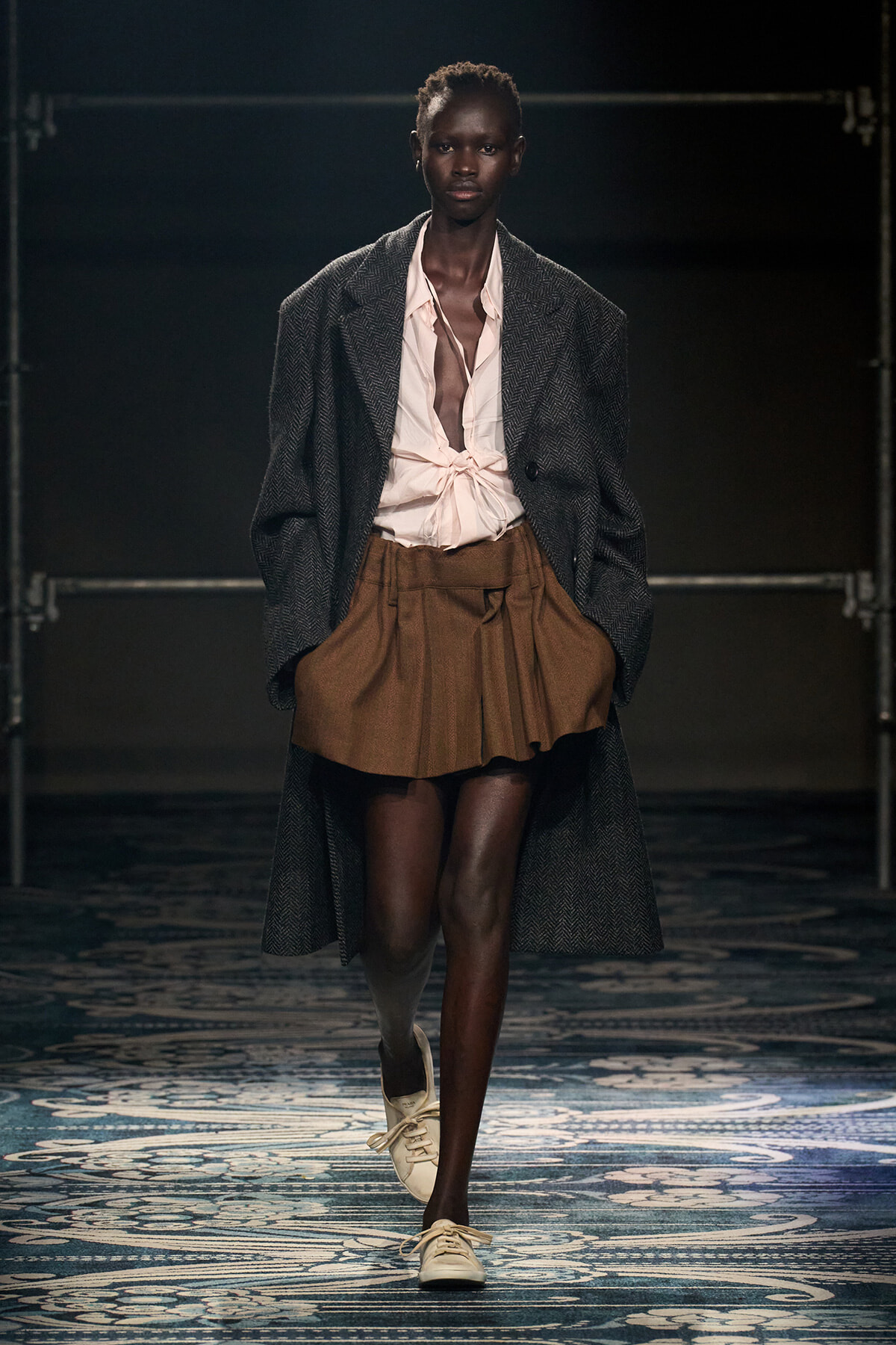 Model walking runway in a dark herringbone overcoat, pink tied blouse, and brown mini skirt; white sneakers visible in a fashion show.