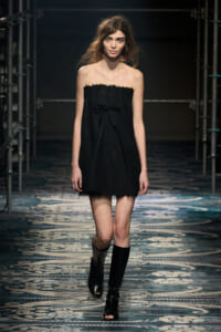 Model walking a fashion show in a strapless black mini dress with a bow, paired with knee-high black boots on a patterned runway floor