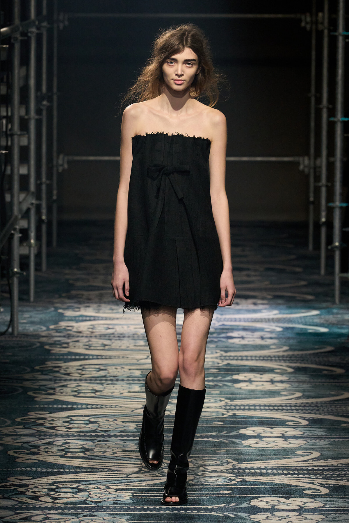 Model walking a fashion show in a strapless black mini dress with a bow, paired with knee-high black boots on a patterned runway floor