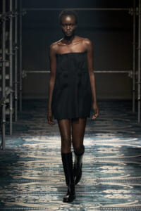 Model walking down a runway in a black strapless mini dress with fringe edges, paired with tall black boots under studio lighting.