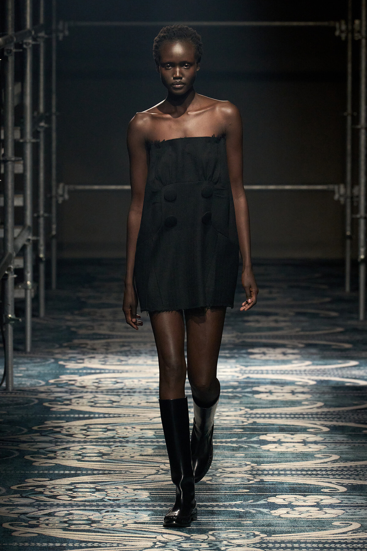 Model walking down a runway in a black strapless mini dress with fringe edges, paired with tall black boots under studio lighting.