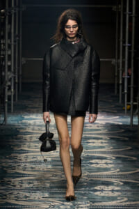 Model walks a runway wearing an oversized black textured coat over a black mini dress, carrying a small black handbag.