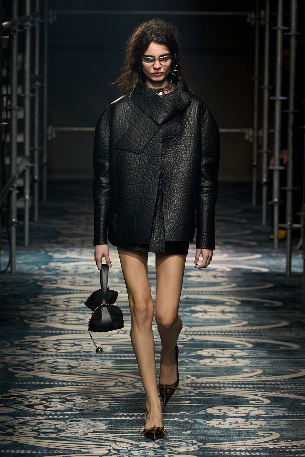 Model walks a runway wearing an oversized black textured coat over a black mini dress, carrying a small black handbag.