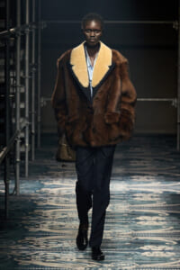 Model walks a runway in a brown fur coat with cream lapels, navy trousers, and a small handbag.