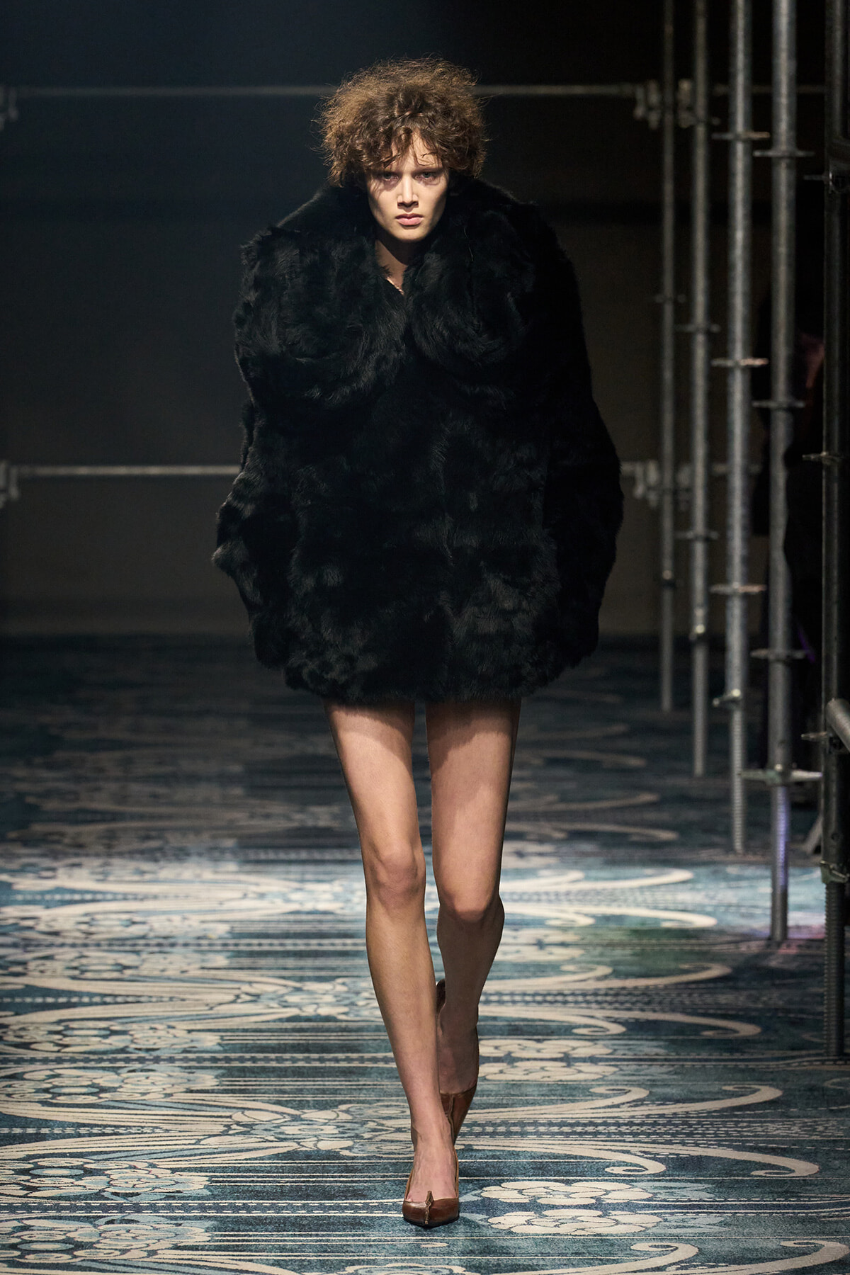 Model walking a runway in a voluminous black fur coat with brown pointed heels on a patterned catwalk.