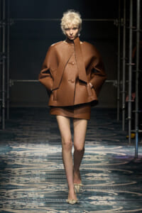 Model on a fashion runway wearing a oversized brown textured coat with wide sleeves and a matching mini skirt; beige heels visible on a patterned catwalk.