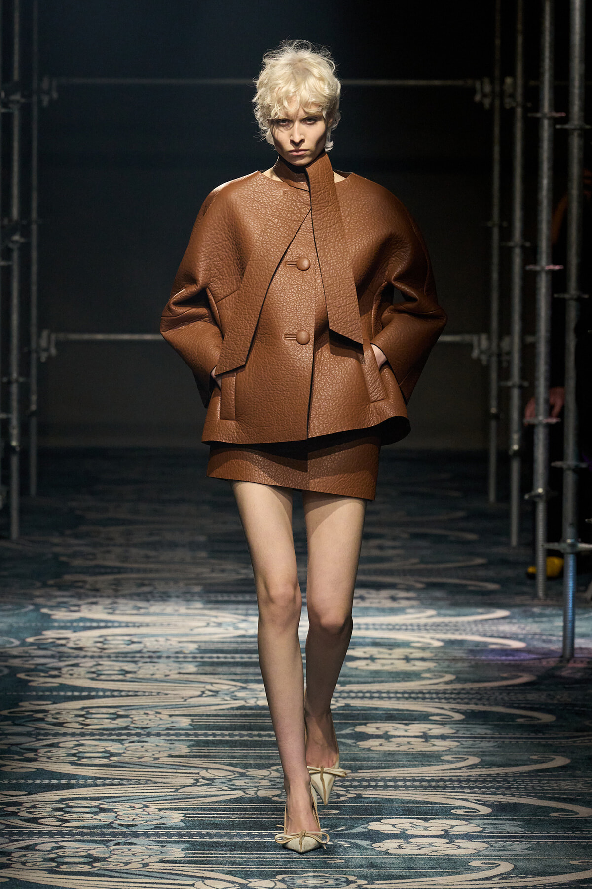 Model on a fashion runway wearing a oversized brown textured coat with wide sleeves and a matching mini skirt; beige heels visible on a patterned catwalk.
