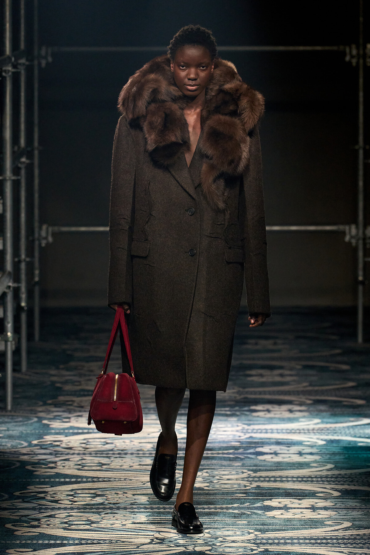 Model walks the runway in a long dark brown coat with a large fur collar, carrying a red handbag and wearing black loafers on a patterned floor.