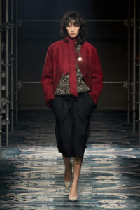 Model on a runway wearing a burgundy oversized jacket over a textured black-and-white top and a black wrap skirt; beige pointed-toe heels.