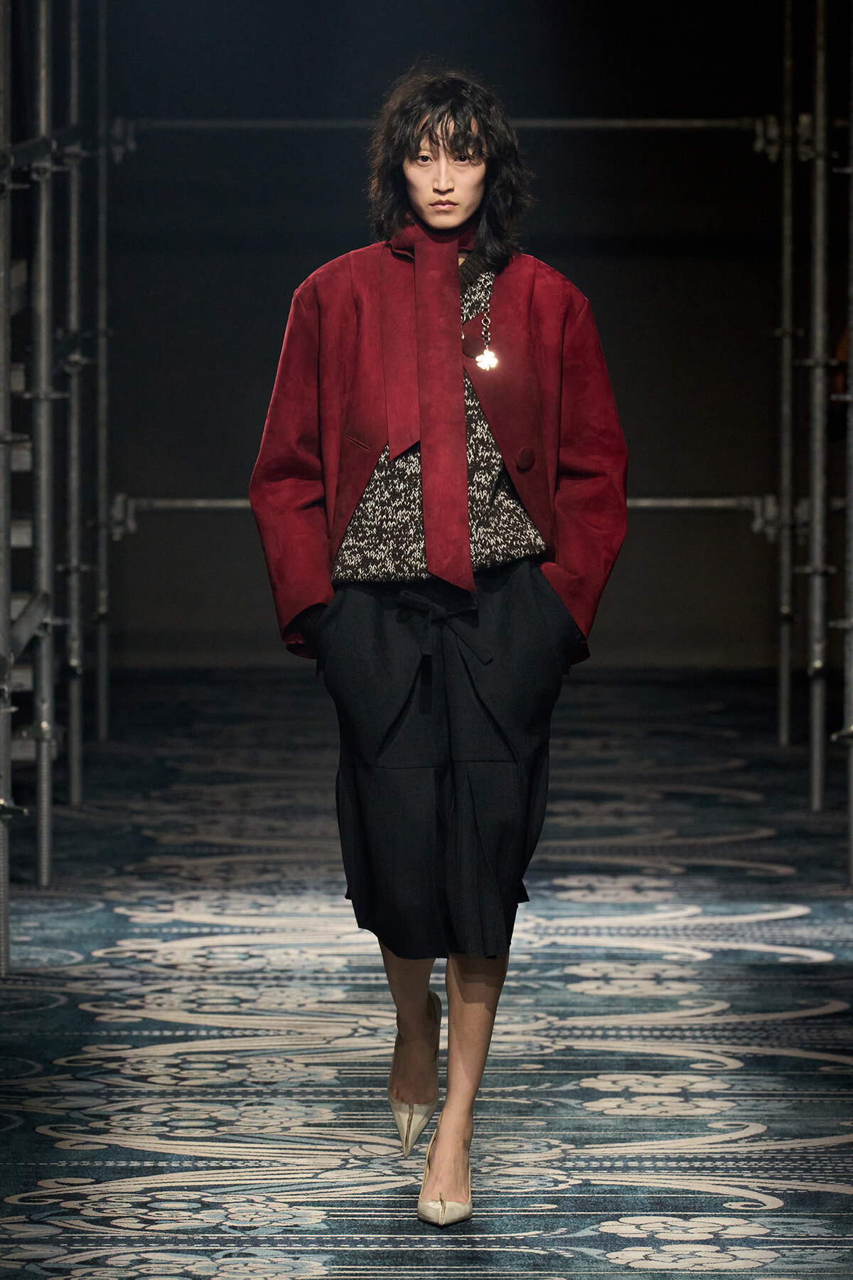 Model on a runway wearing a burgundy oversized jacket over a textured black-and-white top and a black wrap skirt; beige pointed-toe heels.