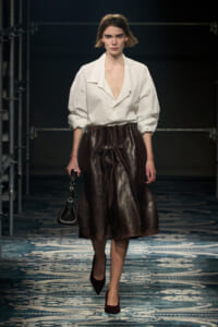 Model walks a fashion show wearing a white oversized blouse with a brown leather knee-length skirt, carrying a small black handbag.