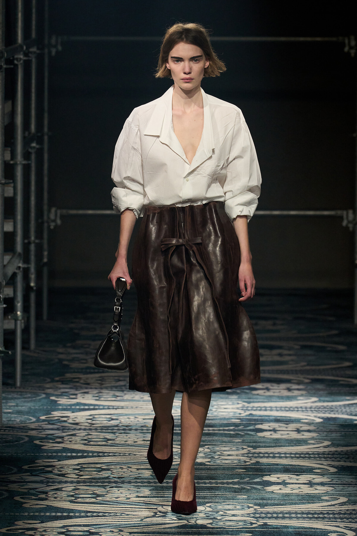 Model walks a fashion show wearing a white oversized blouse with a brown leather knee-length skirt, carrying a small black handbag.