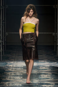 Model walking the runway in a lime yellow bandeau top with white trim and a knee-length brown leather skirt.