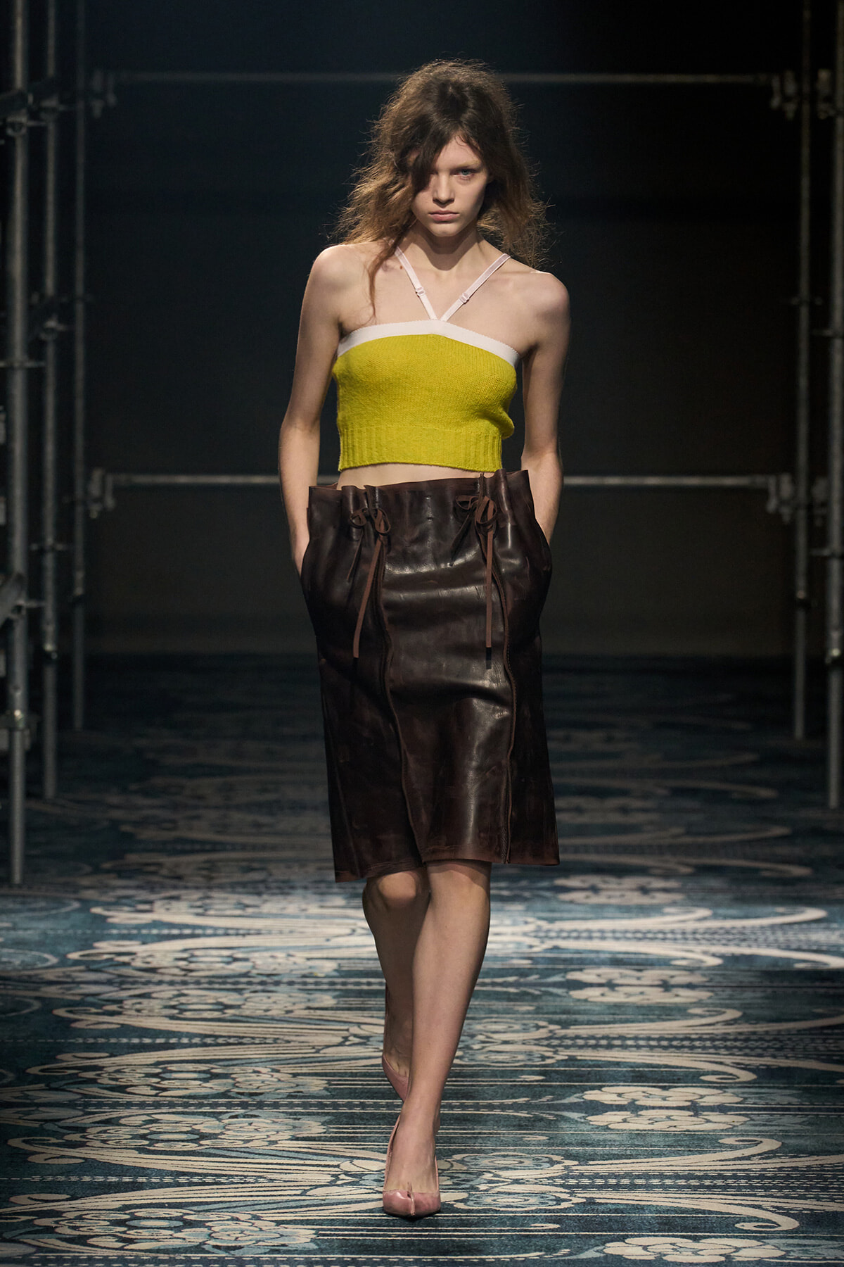 Model walking the runway in a lime yellow bandeau top with white trim and a knee-length brown leather skirt.