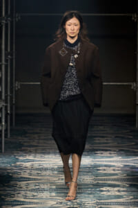 Model walking runway in a dark brown blazer over a patterned knit top, black midi skirt, and silver heels, under dim stage lighting.