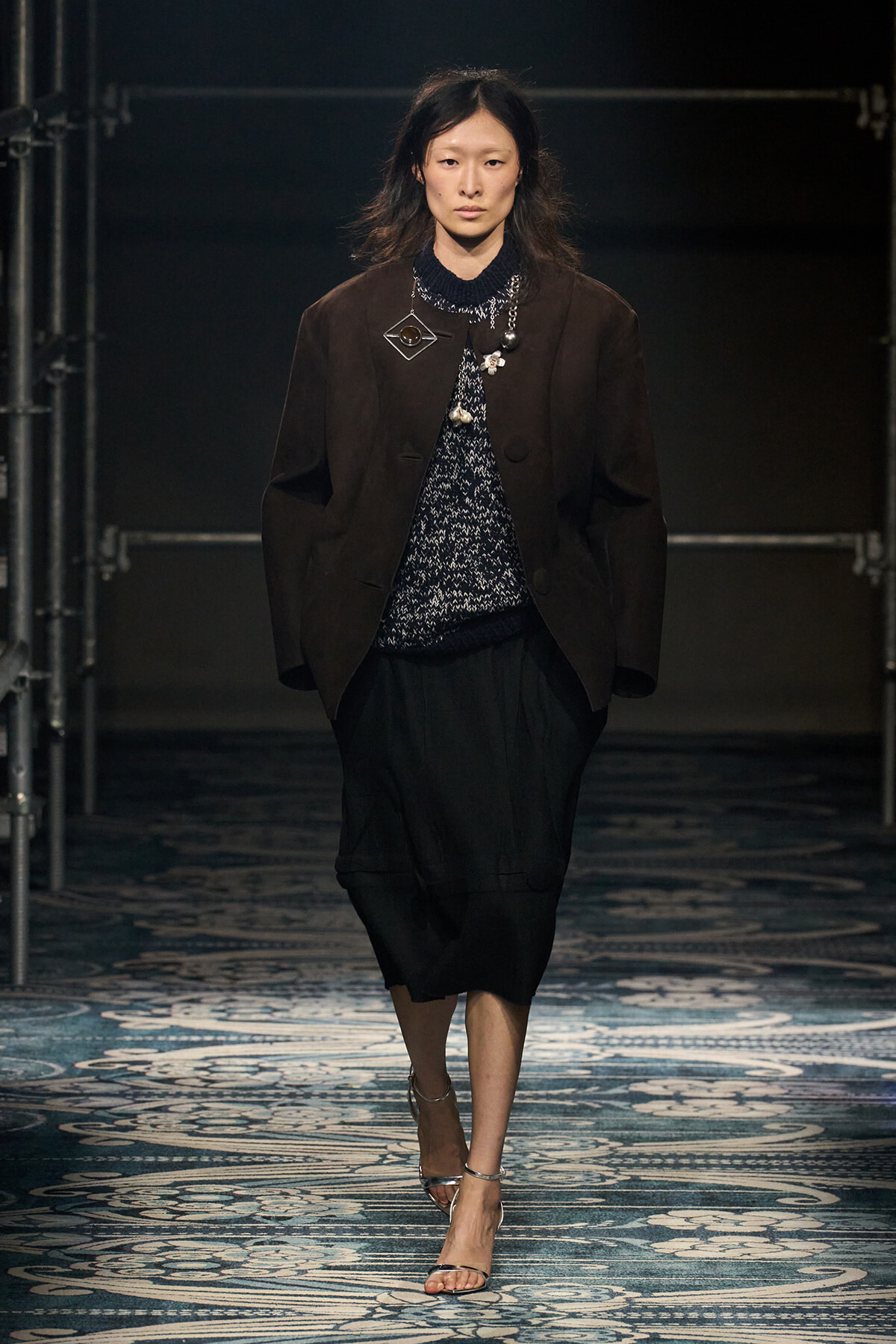 Model walking runway in a dark brown blazer over a patterned knit top, black midi skirt, and silver heels, under dim stage lighting.