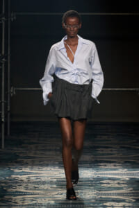 Model walking a runway in a light blue button-down shirt tied at the waist, paired with a gray plaid mini-skirt and black sandals.