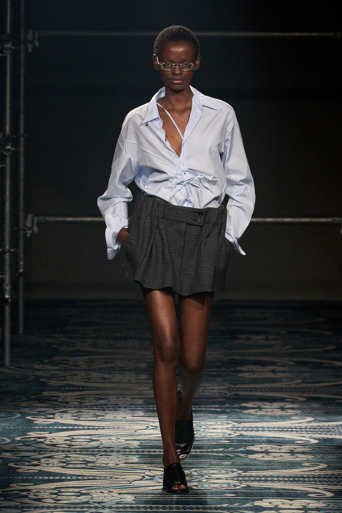 Model walking a runway in a light blue button-down shirt tied at the waist, paired with a gray plaid mini-skirt and black sandals.