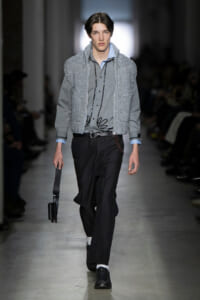 Male model on a runway wearing a gray knitted vest over a gray shirt with black trousers and a small black crossbody bag appearing at his side. Runway look with layered gray knit vest and shirt, black trousers, belt, and polished black shoes. Model walks the fashion show in a pale blue-collar shirt under a chunky gray knit vest, black dress pants, and a black briefcase-style bag. Runway image: gray knit vest over light shirt, dark tailored pants, and a compact black shoulder bag accessory. Elegant menswear look featuring an oversized knit vest paired with tailored trousers and black lace-up shoes.
