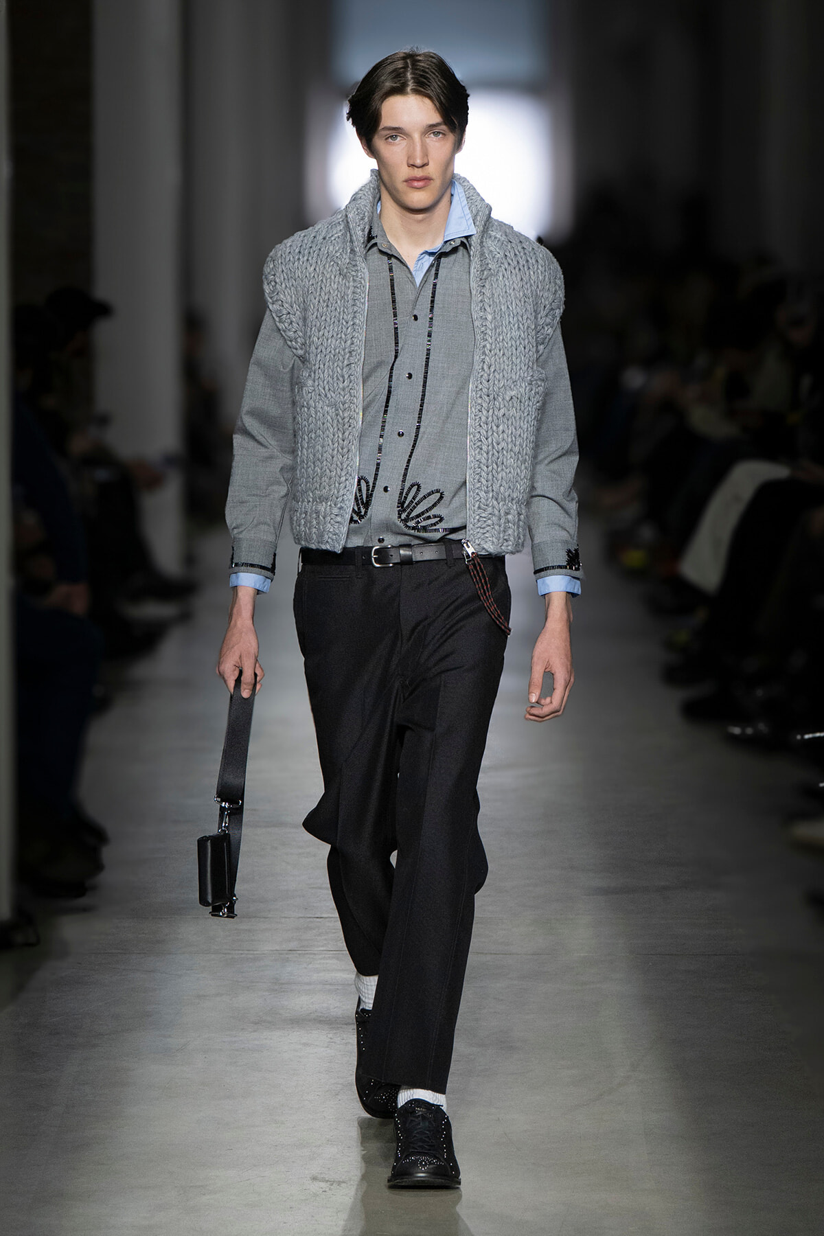 Male model on a runway wearing a gray knitted vest over a gray shirt with black trousers and a small black crossbody bag appearing at his side. Runway look with layered gray knit vest and shirt, black trousers, belt, and polished black shoes. Model walks the fashion show in a pale blue-collar shirt under a chunky gray knit vest, black dress pants, and a black briefcase-style bag. Runway image: gray knit vest over light shirt, dark tailored pants, and a compact black shoulder bag accessory. Elegant menswear look featuring an oversized knit vest paired with tailored trousers and black lace-up shoes.