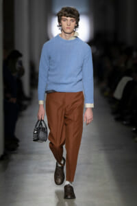 Male model walking a runway in a light blue knit sweater, rust-colored trousers, and dark studded shoes, carrying a small black studded handbag.