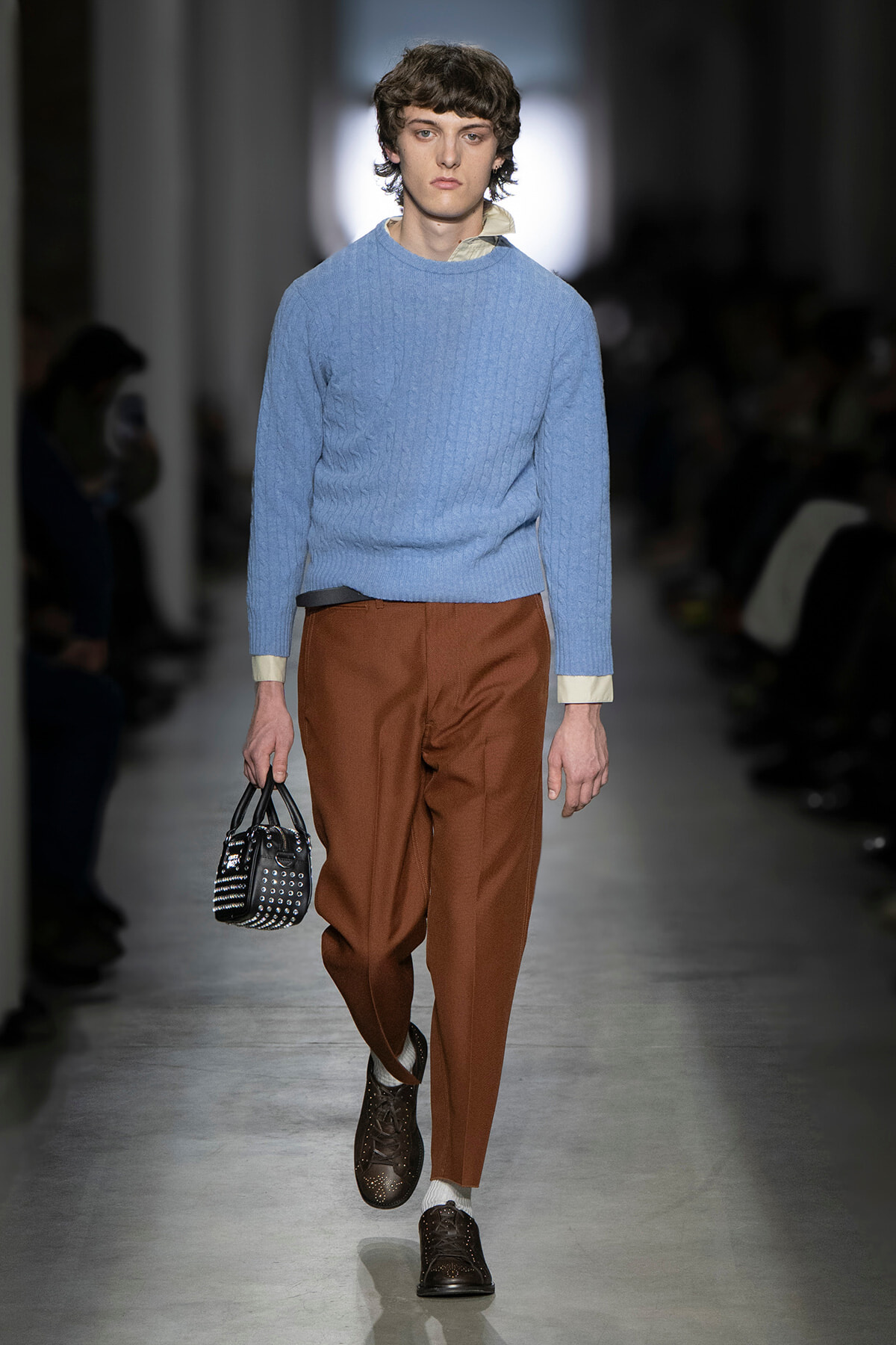 Male model walking a runway in a light blue knit sweater, rust-colored trousers, and dark studded shoes, carrying a small black studded handbag.
