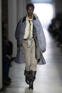 Model walks a fashion runway in a long gray coat over a beige shirt, checkered pants, and black boots with a fringed scarf accessory.