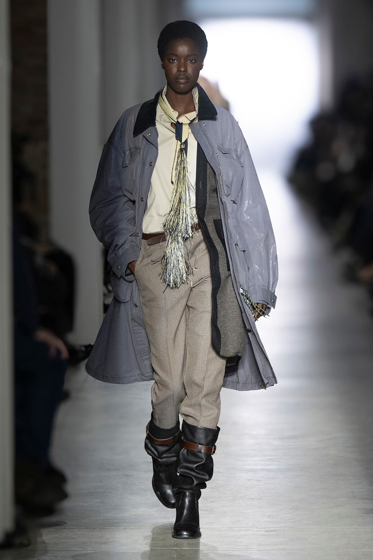 Model walks a fashion runway in a long gray coat over a beige shirt, checkered pants, and black boots with a fringed scarf accessory.