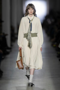 Model on a runway wearing a flowing cream dress with a striped belt, blue fringe necklace, and brown crossbody bag.