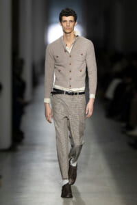 Male model on a runway wearing a light gray knit cardigan with brown buttons, white shirt, and beige plaid trousers with a black belt and brown brogue shoes.