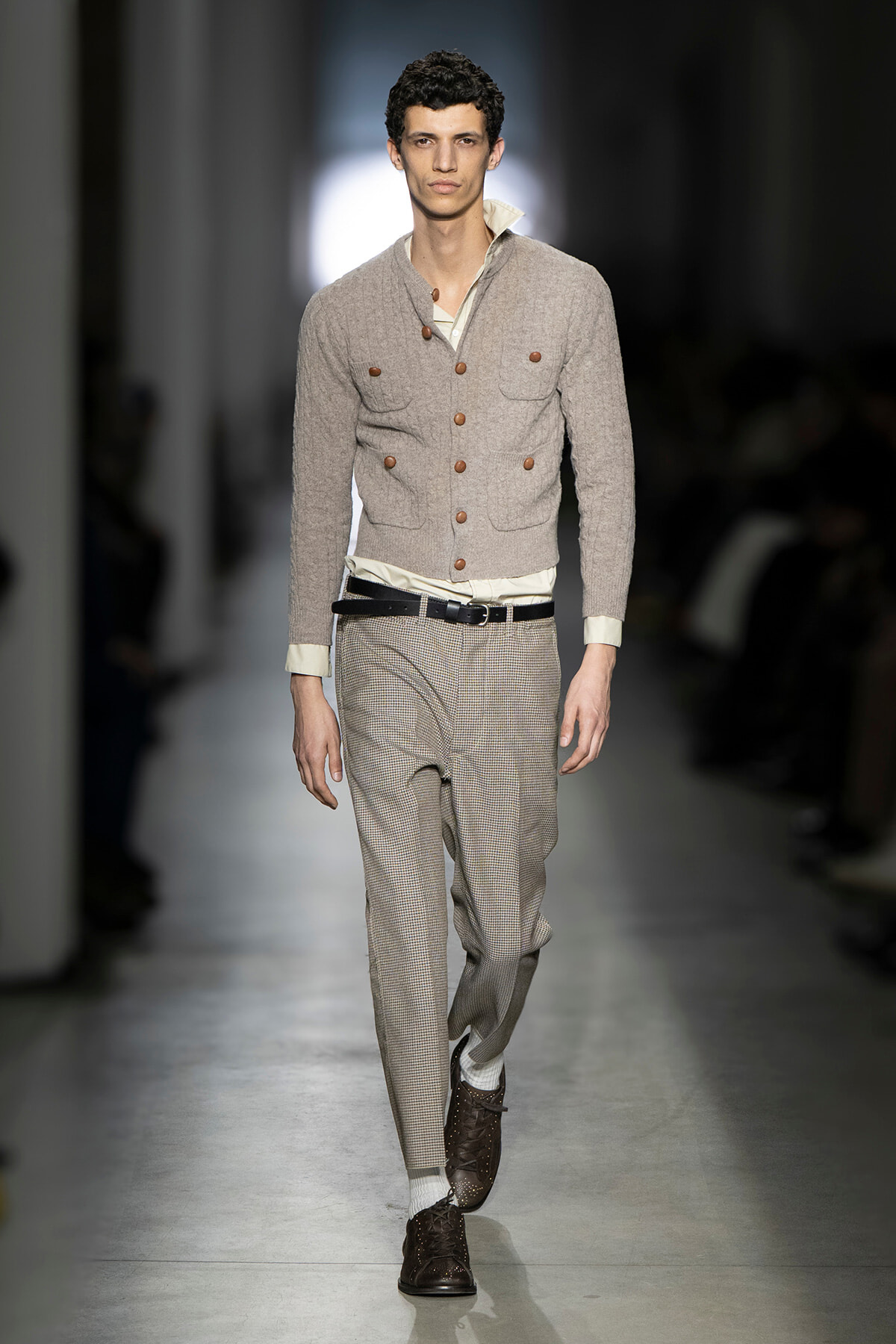 Male model on a runway wearing a light gray knit cardigan with brown buttons, white shirt, and beige plaid trousers with a black belt and brown brogue shoes.