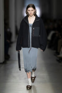 Model walking a runway in an oversized black zip-front jacket over a blue-gray dress, white socks, and brown sandals, holding a black strap bag edgewise.