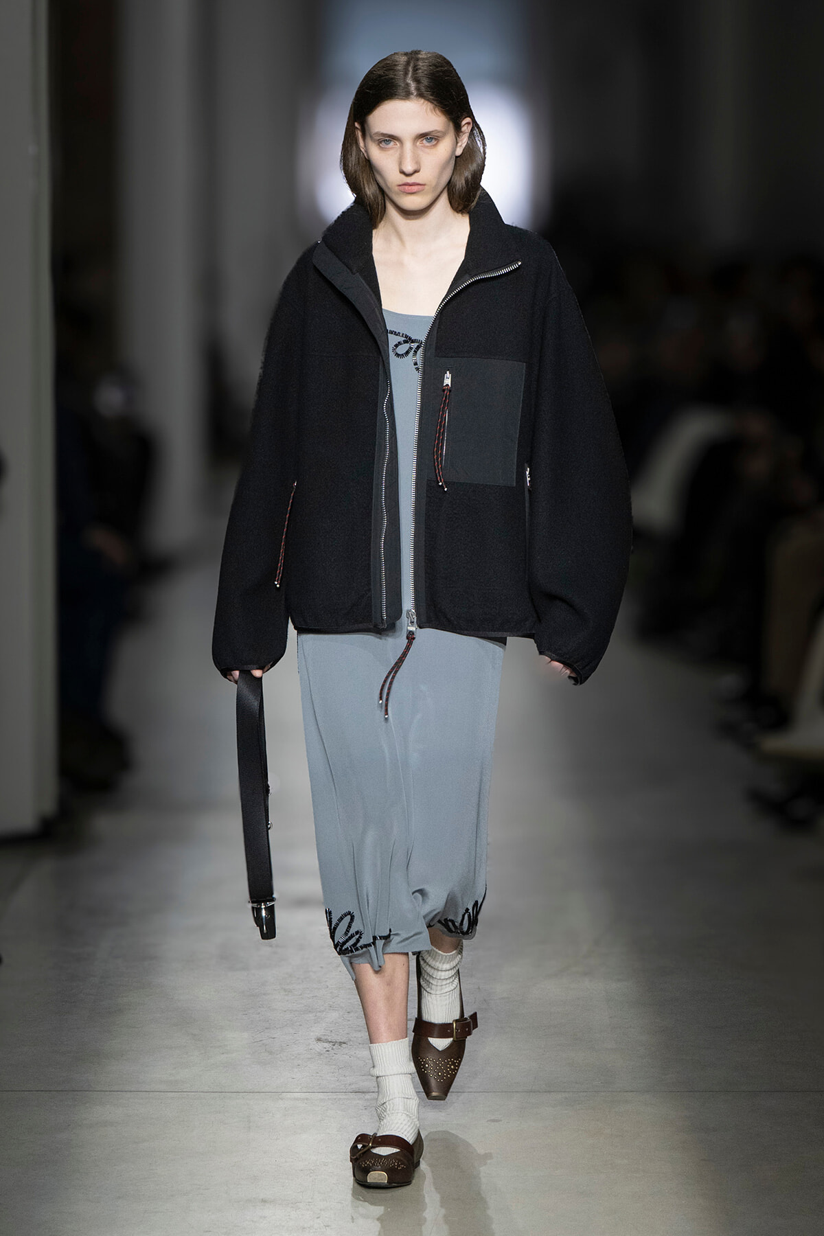 Model walking a runway in an oversized black zip-front jacket over a blue-gray dress, white socks, and brown sandals, holding a black strap bag edgewise.