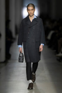 Male model walks a runway in a dark double-breasted wool coat with a light blue shirt peeking at the collar, carrying a studded black handbag.