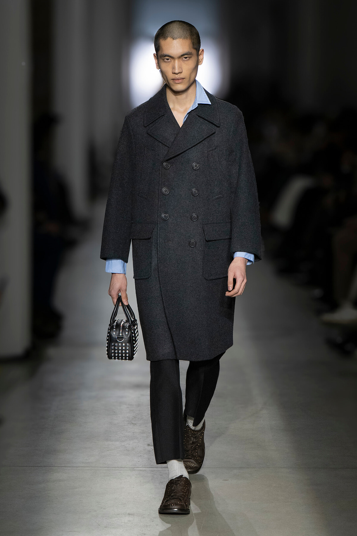 Male model walks a runway in a dark double-breasted wool coat with a light blue shirt peeking at the collar, carrying a studded black handbag.