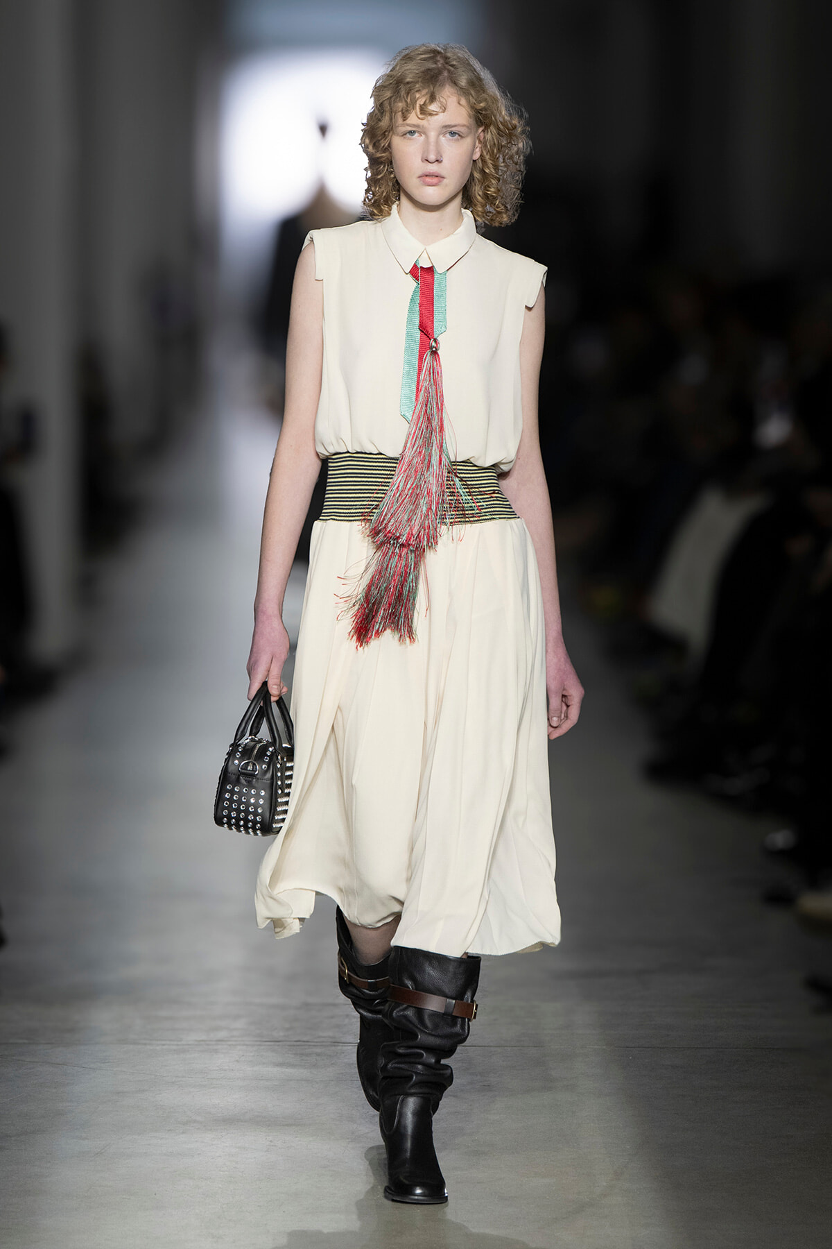 Model on runway wearing a cream sleeveless dress with a wide striped belt and colorful tassel, carrying a studded black handbag and wearing black boots.