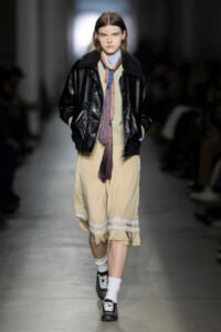 Model on a runway wearing a black leather bomber with shearling, a beige midi dress with lace trim, a colorful scarf, white socks, and studded black shoes.