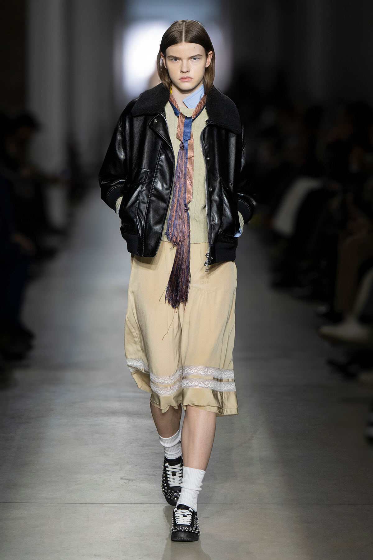 Model on a runway wearing a black leather bomber with shearling, a beige midi dress with lace trim, a colorful scarf, white socks, and studded black shoes.