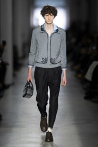 Male model walking a runway in a light-gray zipped jacket with black embroidery, pale blue shirt, black trousers, and brown brogue shoes, carrying a studded black handbag.