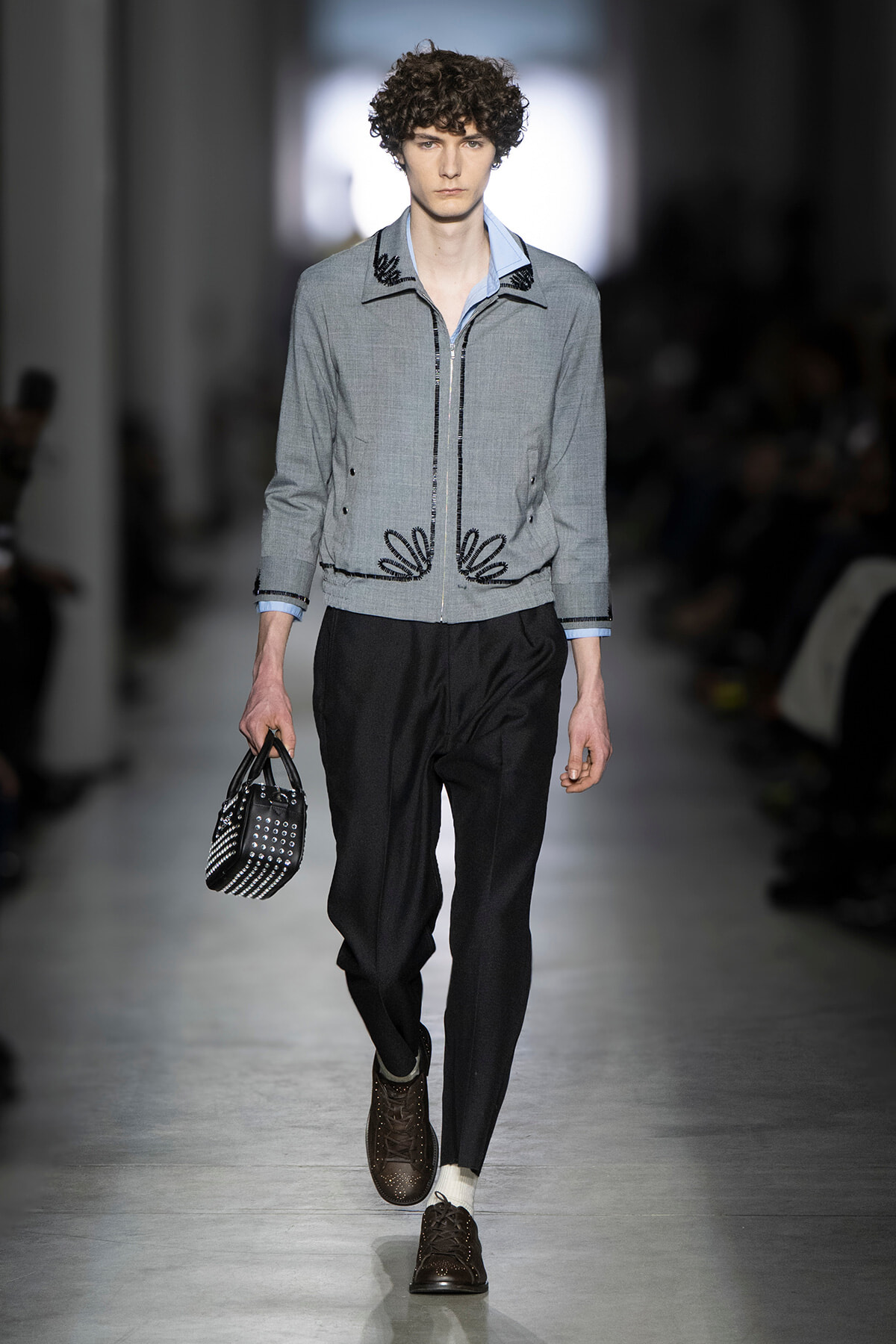 Male model walking a runway in a light-gray zipped jacket with black embroidery, pale blue shirt, black trousers, and brown brogue shoes, carrying a studded black handbag.