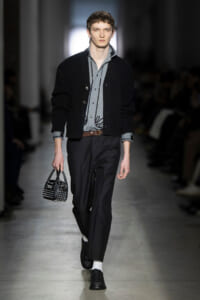 Male model walks the runway in a black cardigan over a gray button-down shirt, dark trousers, brown belt, and a black studded handbag.