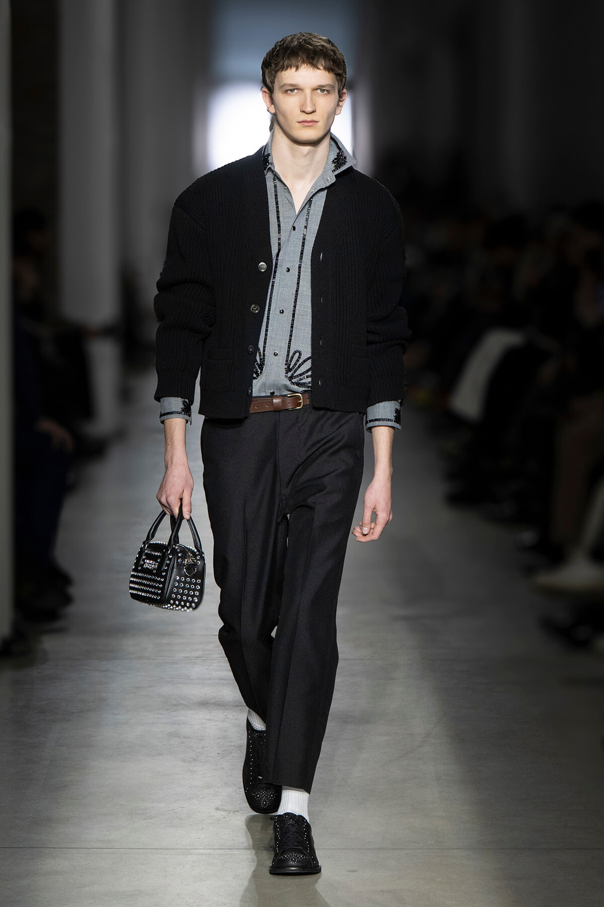 Male model walks the runway in a black cardigan over a gray button-down shirt, dark trousers, brown belt, and a black studded handbag.