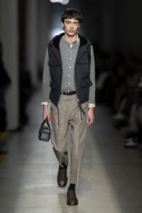 Male model walks the runway in a gray layered outfit: knit gray shirt, dark hooded vest, beige checkered trousers, brown brogues, and a studded black handbag accessory moved in hand.