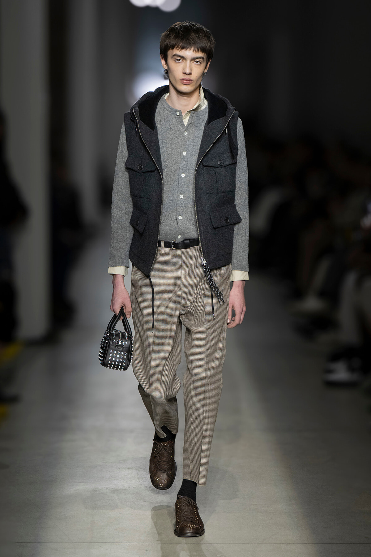 Male model walks the runway in a gray layered outfit: knit gray shirt, dark hooded vest, beige checkered trousers, brown brogues, and a studded black handbag accessory moved in hand.