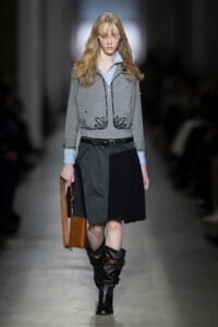 Runway model in a gray embroidered jacket over a light blue shirt, with a color-block skirt, black knee-high boots, and a brown bag.
