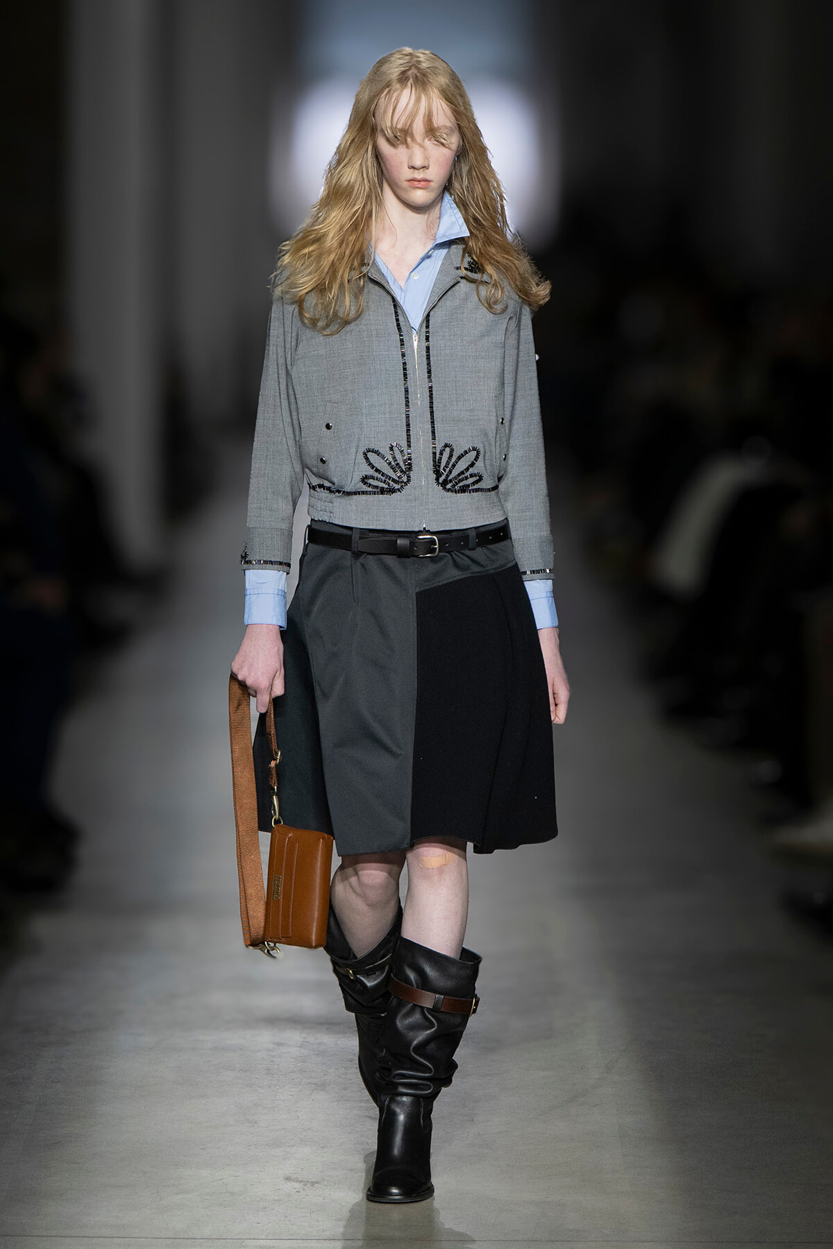 Runway model in a gray embroidered jacket over a light blue shirt, with a color-block skirt, black knee-high boots, and a brown bag.